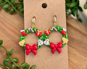 May include: A pair of festive Christmas wreath earrings. Each earring features a green wreath adorned with red bows, candy canes, gift boxes, snowmen, and gold bells. The earrings are attached to gold-colored ear wires and displayed on a brown card.