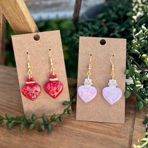 May include: Two pairs of heart-shaped earrings. One pair is red with gold-coloured accents, and the other is a light pink with glitter. Both pairs have gold-coloured ear wires and are displayed on brown card.