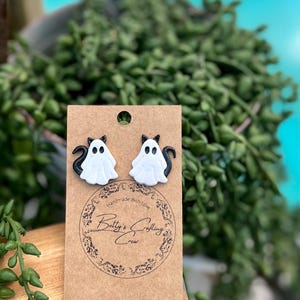 Op de afbeelding: Een paar zwarte en witte spookoorbellen met een zwart kattengezicht op elke spook. De oorbellen bevinden zich op een bruine kaart met de tekst "Handmade with love Betty's Crafting Crew".
