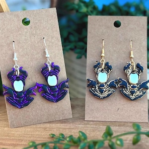 May include: Two pairs of handmade earrings featuring a stylized character design. One pair is purple and black with silver hooks, the other is black and gold with gold hooks. Each earring has a light green center.