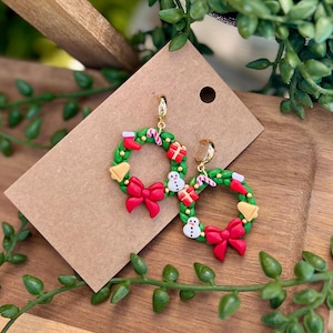 May include: A pair of festive Christmas wreath earrings. Each earring features a green wreath adorned with red bows, gift boxes, snowmen, candy canes, and a yellow bell. The earrings have gold-colored hoops and are set against a brown card.