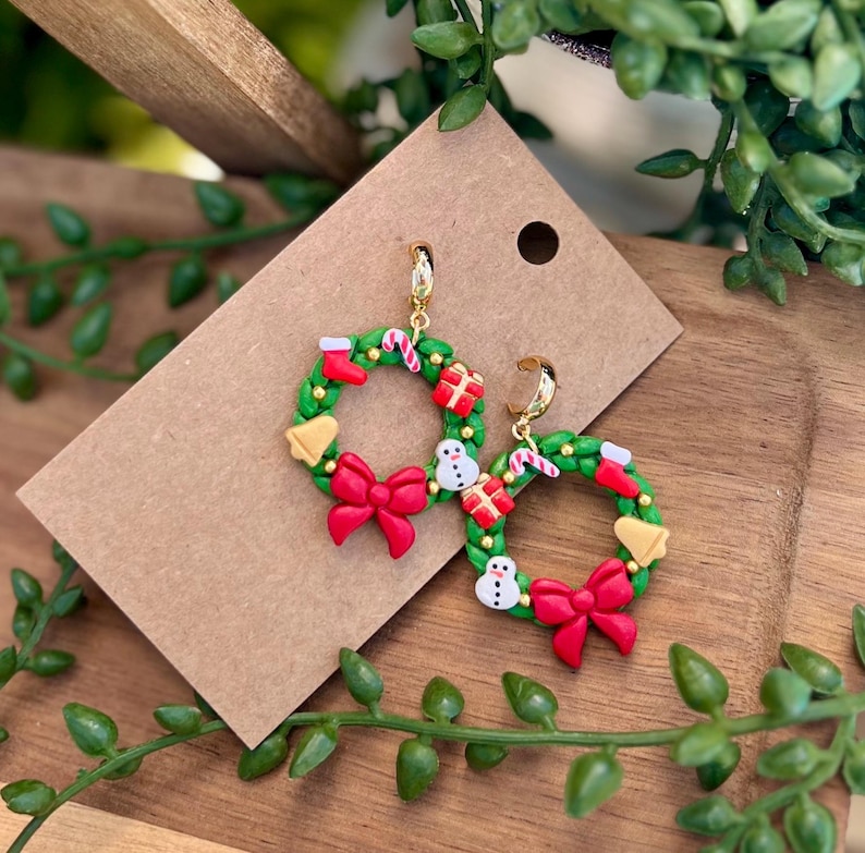 May include: A pair of festive Christmas wreath earrings. Each earring features a green wreath adorned with red bows, gift boxes, snowmen, candy canes, and a yellow bell. The earrings have gold-colored hoops and are set against a brown card.