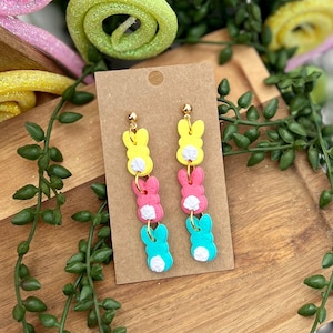 Puede incluir: Un par de pendientes colgantes con tres abalorios de conejito en colores pastel. Los abalorios son amarillos, rosas y azules con detalles blancos. Los pendientes están en una tarjeta marrón.