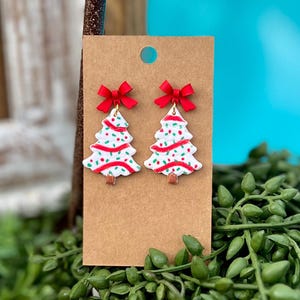 Puede incluir: Un par de pendientes de árbol de Navidad. Cada pendiente presenta una forma de árbol blanco con detalles rojos y verdes, rematado con un lazo rojo. Los pendientes se exhiben en una tarjeta marrón.