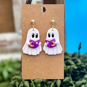 Peut inclure: Une paire de boucles d'oreilles pendantes en forme de fant&ocirc;me blanc avec des yeux noirs, tenant un livre violet orn&eacute; d'un croissant de lune et d'une &eacute;toile dor&eacute;s. Elles sont fix&eacute;es sur un support en carton brun avec des clous dor&eacute;s.