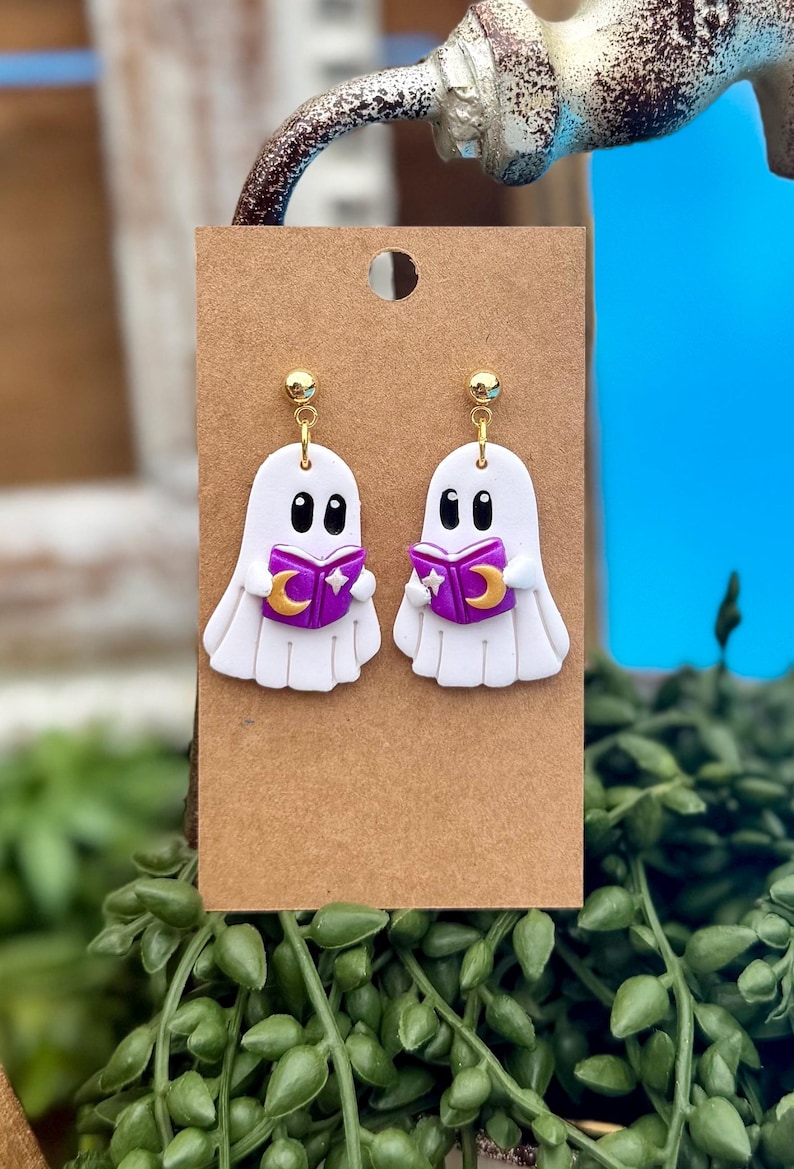 Peut inclure: Une paire de boucles d'oreilles pendantes en forme de fant&ocirc;me blanc avec des yeux noirs, tenant un livre violet orn&eacute; d'un croissant de lune et d'une &eacute;toile dor&eacute;s. Elles sont fix&eacute;es sur un support en carton brun avec des clous dor&eacute;s.