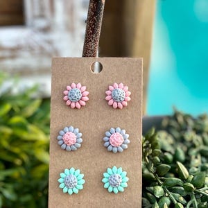 May include: A set of six stud earrings featuring three different flower designs. The flowers are pink, gray, and mint green with a gray center. The earrings are displayed on a brown card.