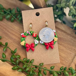 May include: A pair of Christmas wreath earrings. Each earring features a green wreath with red bow, candy cane, gift, snowman, and gold bell accents. The earrings are displayed on a brown card.