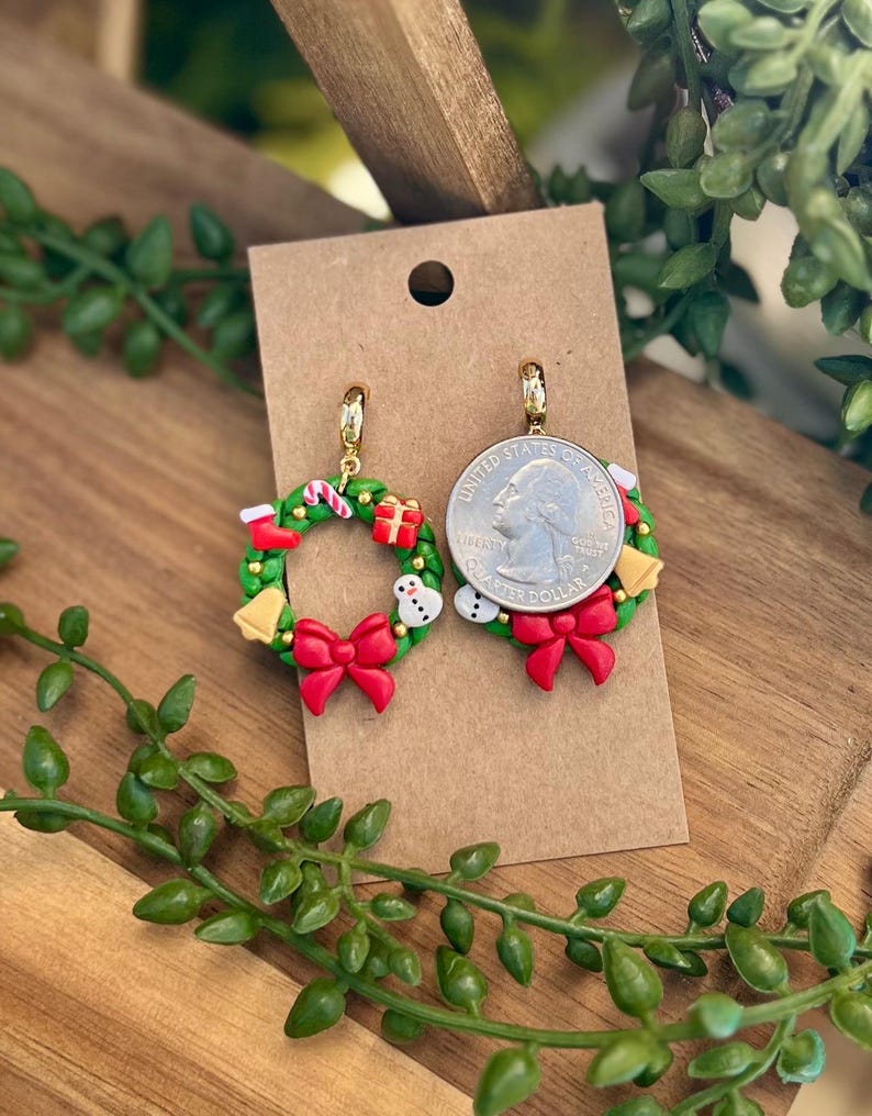 May include: A pair of Christmas wreath earrings. Each earring features a green wreath with red bow, candy cane, gift, snowman, and gold bell accents. The earrings are displayed on a brown card.