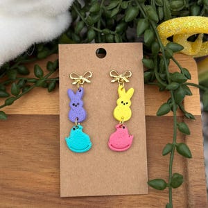 May include: Colorful Easter-themed earrings. Each earring features a gold bow at the top, with a dangling design of a purple bunny, a turquoise chick, a yellow bunny, and a pink chick. The earrings are displayed on a brown card.