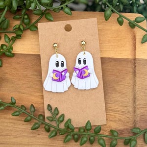 Peut inclure: Une paire de boucles d'oreilles en forme de fant&ocirc;me blanc avec des yeux noirs, tenant chacune un livre violet orn&eacute; d'un croissant de lune jaune et d'une &eacute;toile blanche. Elles sont fix&eacute;es sur des tiges dor&eacute;es et pr&eacute;sent&eacute;es sur un carton brun.