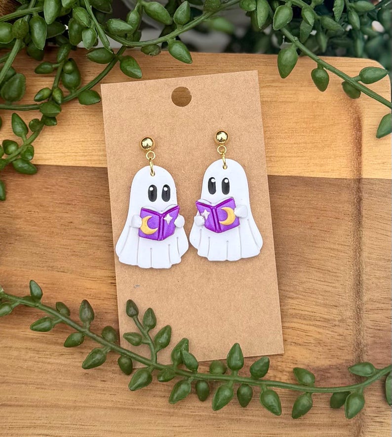 Peut inclure: Une paire de boucles d'oreilles en forme de fant&ocirc;me blanc avec des yeux noirs, tenant chacune un livre violet orn&eacute; d'un croissant de lune jaune et d'une &eacute;toile blanche. Elles sont fix&eacute;es sur des tiges dor&eacute;es et pr&eacute;sent&eacute;es sur un carton brun.