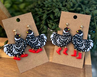 May include: Two pairs of dangle earrings shaped like chickens. Each chicken is black with white polka dots and stripes, featuring a small red comb, a yellow beak, and bright red boots. The earrings are attached to gold-tone posts.