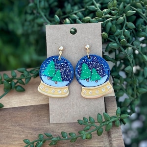 May include: A pair of dangle earrings featuring a snow globe design with a dark blue glittery sky, two green pine trees, and a white snowy base. The base of the snow globe is light yellow with white star accents and gold-tone hardware.