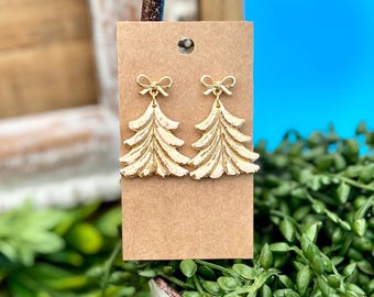 May include: Gold-colored Christmas tree earrings with a bow at the top. The earrings are displayed on a brown card with a hole at the top. The background includes greenery and a blue surface.