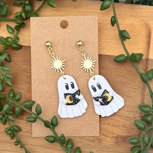 Peut inclure: Une paire de boucles d'oreilles pendantes avec des breloques fant&ocirc;mes blanches tenant des livres noirs orn&eacute;s d'un croissant de lune jaune et d'&eacute;toiles blanches. Chaque fant&ocirc;me est suspendu &agrave; un connecteur soleil dor&eacute; sur un support en carton brun.