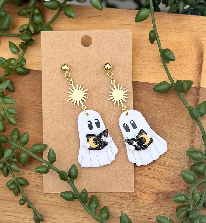 Peut inclure: Une paire de boucles d'oreilles pendantes avec des breloques fant&ocirc;mes blanches tenant des livres noirs orn&eacute;s d'un croissant de lune jaune et d'&eacute;toiles blanches. Chaque fant&ocirc;me est suspendu &agrave; un connecteur soleil dor&eacute; sur un support en carton brun.