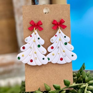 May include: A pair of white Christmas tree-shaped earrings featuring red bows at the top. The trees are decorated with small, embedded red, green, and clear rhinestones along with tiny gold-colored spheres, all displayed on a brown card.