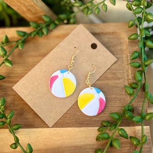 May include: A pair of beach ball earrings with gold-colored hooks. Each earring features a white circle with segments of yellow, pink, and blue. The earrings are displayed on a brown card against a wooden background, with green foliage.