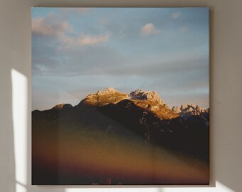 May include: A framed photograph of a mountain range with a cloudy sky in the background. The mountain range is lit by the sun, creating a warm glow. The photograph is hanging on a white wall.