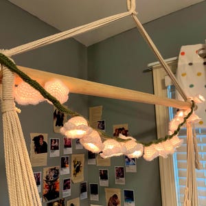 May include: A hanging macrame swing with a wooden bar, adorned with a string of illuminated, crocheted flower lights. The lights are white and attached to a green crocheted vine. The swing is suspended from the ceiling.