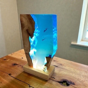 Könnte beinhalten: Eine rechteckige Lampe mit einem vertikalen Stück Naturholz mit Baumkante, kombiniert mit durchscheinendem blauem Harz, das tiefes Wasser nachahmt. Zwei kleine Tauchersilhouetten sind im Harz eingebettet und werden von der Basis beleuchtet.