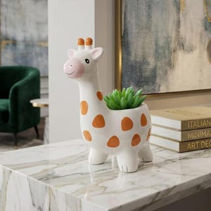 May include: A white ceramic giraffe planter with orange spots, holding a green succulent. The giraffe has a pink nose and ears. The planter is on a marble surface, with books and artwork in the background.