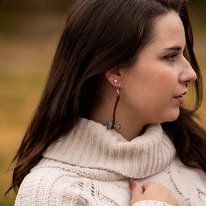 May include: Axe-shaped earrings with silver-colored axe heads and brown leather-like handles. The earrings are suspended from silver-colored hooks. The background is blurred, with a person wearing a cream-colored sweater.