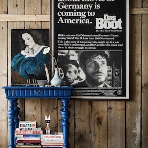May include: A royal blue side table with detailed legs supports books and ornaments. A framed movie poster with the text "Das Boot" is displayed on a wooden wall, next to a portrait painting.