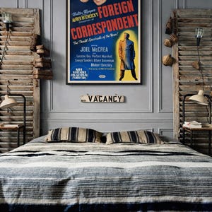 May include: A framed vintage movie poster for Alfred Hitchcock's Foreign Correspondent featuring a blue background, red text, and illustrations of the cast. Below the poster is a rectangular wooden sign with the text NO VACANCY in black letters.
