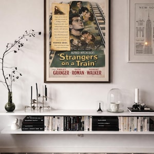 May include: A framed vintage movie poster for Alfred Hitchcock's Strangers on a Train, featuring illustrations of the cast and train tracks, displayed above a white wall-mounted shelf containing books, a glass cloche, and a metal candle holder.