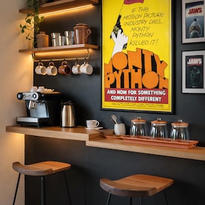 Könnte beinhalten: Eine Kaffeebar mit zwei Holzhockern auf schwarzen Metallbeinen, einer Holztheke mit Espressomaschine, Wasserkocher und drei Glasbehältern. Zwei Holzregale beherbergen Pflanzen, Tassen und ein gerahmtes Monty Python Filmplakat.