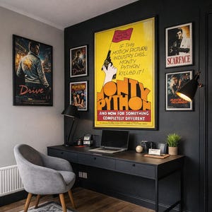 May include: A home office setup featuring a black desk with a laptop, a gray upholstered chair, and a large Monty Python movie poster on a dark wall. Surrounding posters include Drive, Pulp Fiction, Scarface, and Blade Runner, with a floor lamp.
