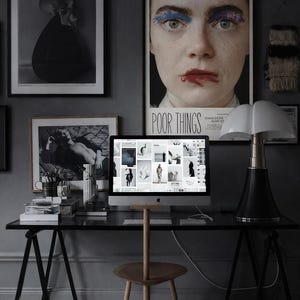 May include: A black desk with a computer monitor displaying a grid of images, a wooden three-legged stool, and a black and white table lamp. The wall features framed art prints, including a large poster with the text Poor Things.