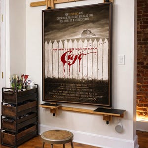 May include: A framed movie poster with the title "Cujo" hangs on a white wall. The poster is mounted on a wooden frame with two small shelves. A teal-colored wall lamp is above the poster. A metal stool sits on the wooden floor.