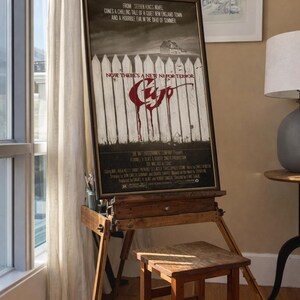 May include: A framed Cujo movie poster featuring a white picket fence, a house in the distance, and the red dripping title text. The poster includes the text: From Stephen King's novel comes a chilling tale of a quiet New England town.