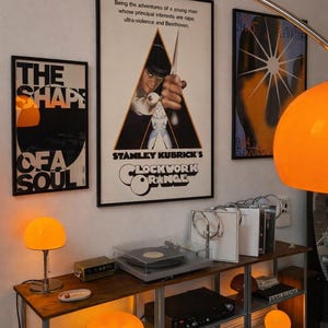 Op de afbeelding: Een metalen rek met een platenspeler, vinylplaten en oranje lampen. Aan de muur hangen drie ingelijste posters, waaronder een centrale A Clockwork Orange filmposter met tekst en een afbeelding van een persoon die een mes vasthoudt.