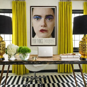 May include: A home office with a wooden desk, a laptop, and a framed poster with the text "POOR THINGS". The room features yellow curtains, black lamps, and a black and white striped rug. Decorative flowers and a white office chair are also present.