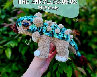 May include: A crocheted ankylosaurus dinosaur toy in shades of tan, teal, and brown. The toy has a textured body, a patterned back, and small, round spikes. The text "Aspen THE ANKYLOSAURUS PDF Only" is displayed above the toy. The words "Crochet Pattern" are displayed below the toy.