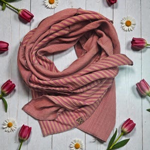 Könnte beinhalten: Ein rosa-beiger gestreifter Schal, drapiert und präsentiert. Rote Tulpen und weiße Gänseblümchen umgeben den Schal auf einer weißen Holzoberfläche. Ein kleines Etikett mit Leopardenmuster ist sichtbar.