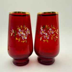 Pode incluir: Dois vasos de vidro vermelho com bordas douradas e desenhos florais. Cada vaso apresenta um grupo de flores brancas e rosas com detalhes dourados. Os vasos têm uma base arredondada e um topo ligeiramente alargado.