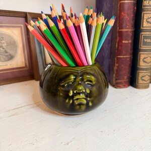 Pode incluir: Um porta-lápis de cerâmica verde escuro com um design de rosto, cheio de lápis de cor. Os lápis estão dispostos em forma de leque, mostrando suas cores vibrantes. O porta-lápis está sobre uma superfície branca.