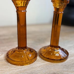 May include: Two amber-colored glass candle holders. The candle holders have a round base with a decorative pattern, a long, slender stem, and a flared top. The glass has a glossy finish, reflecting light. The candle holders are a decorative home accent.