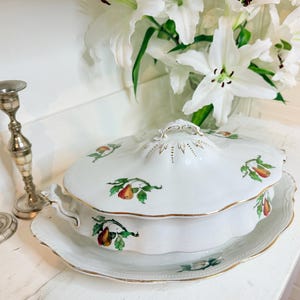 Puede incluir: Fuente de cerámica blanca con tapa, con diseño de pera y ribete dorado. Dos candelabros plateados con detalles en color marfil a la izquierda. Un ramo de lirios blancos en el fondo.