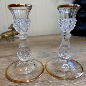 May include: Two clear glass candle holders with gold rims. The candle holders have a diamond-cut pattern on the stem and a round base. The top of the candle holders also has a gold rim. The candle holders are on a wooden surface.