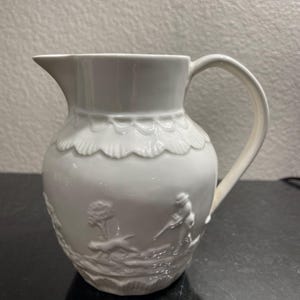 Może przedstawiać: Biały dzbanek ceramiczny z wypukłym wzorem przedstawiającym myśliwego i królika. Dzbanek ma falisty brzeg i uchwyt.