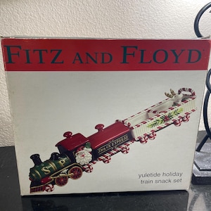 May include: A red, green and white ceramic train snack set with a Santa Claus figure on the engine. The box reads "Fitz and Floyd" and "Yuletide Holiday Train Snack Set".