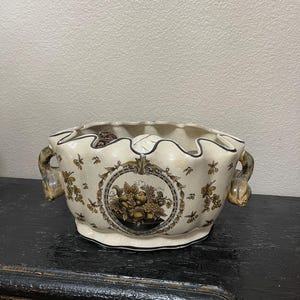 May include: Cream-colored ceramic bowl with a scalloped rim and two ornate handles. The bowl features a central medallion with a fruit and foliage design, and is decorated with small floral motifs.