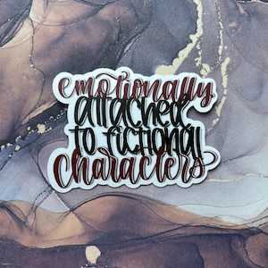 May include: A white sticker with the text "emotionally attached to fictional characters" in black and burgundy script. The sticker has a white border and is set against a marbled background with brown, gold, and white tones.