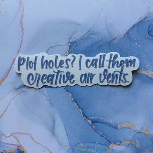 May include: A white sticker with blue text that reads "Plot holes? I call them creative air vents." The sticker is in a cloud shape and is set against a blue and gold marbled background.